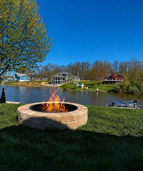 A round stone fire pit burning on a trimmed lawn beside a lake.