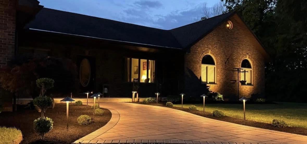 A brick home at dusk with a lit paver walkway, path lights, and fresh mulch beds.